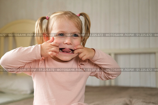 Little Girl joking sticking tongue out with silly expression. Child making face. Funny foolishness portrait of baby girl laughing in bedroom. Adorable Caucasian kid doing crazy comical gesture at home. Стоковое фото, фотограф Марина Демидюк / Фотобанк Лори