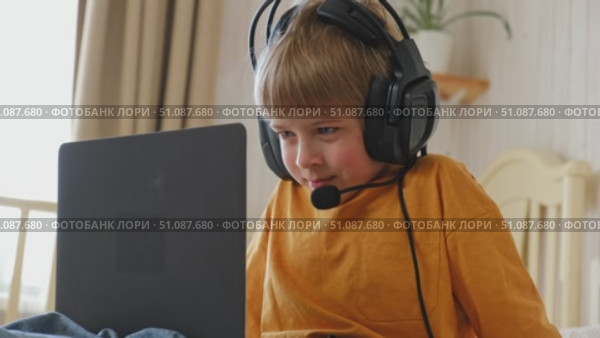 Little smiling 6 years old boy using a laptop with headphones. Child having video call with friends, granny at home in bed. Kid looking at display and waving hand. Childhood leisure activity concept. Стоковое видео, видеограф Марина Демидюк / Фотобанк Лори