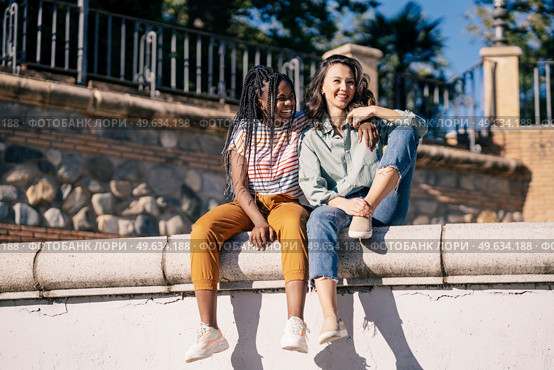 Low angle of cheerful multiethnic female best friends cuddling and sitting on concrete border in street while looking away. Positive diverse women sitting on fence in city. Стоковое фото, агентство Ingram Publishing / Фотобанк Лори