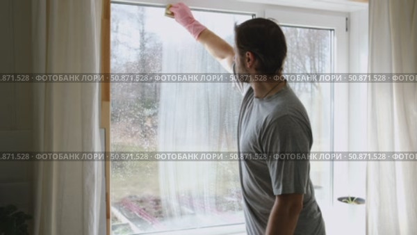 Man is cleaning window in apartment. Adult Male cleaner using a squeegee to wash a window. Bearded guy wiping and washing glasses with with spray bottle, rag and vacoom. Everyday chores. Purification. Стоковое видео, видеограф Марина Демидюк / Фотобанк Лори