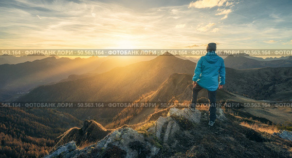 Man on stone on the hill and beautiful mountain valley in haze at colorful sunset in autumn. Dolomites, Italy. Guy, mountain ridges in fog, orange grass and trees, blue sky with sun in fall. Hiking	. Стоковое фото, агентство Ingram Publishing / Фотобанк Лори