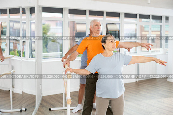 Mature woman in sportswear participating 2nd position of ballet stance at barre with group in choreography studio. Стоковое фото, фотограф Яков Филимонов / Фотобанк Лори