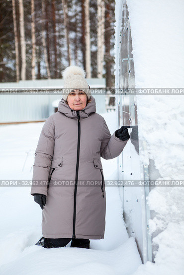 Mature woman standing in snowdrift next to door of her plastic greenhouse on backyard of country house. Стоковое фото, фотограф Кекяляйнен Андрей / Фотобанк Лори