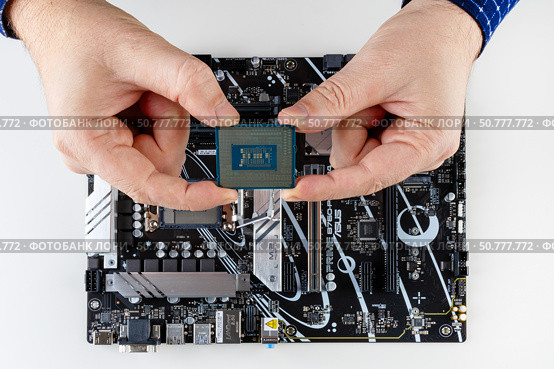 Maykop, Russia - February 18, 2025: men's hands hold the new Intel processor above the Asus motherboard on a light gray background, top view. Редакционное фото, фотограф Андрей С / Фотобанк Лори