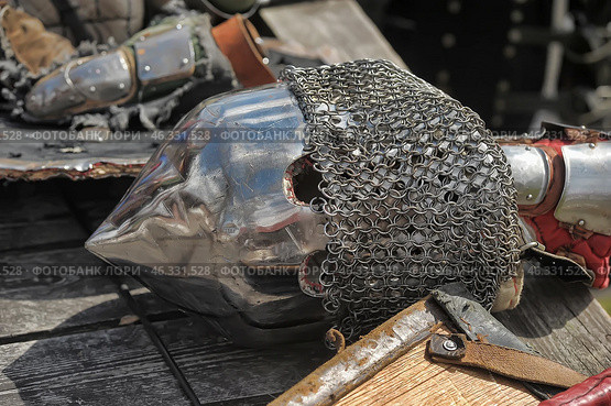 Medieval knight's helmet - bascinet with aventail (chainmail protection for the neck). The helmet is made of metal, polished to a shine, but has traces of scratches and abrasions, which indicates its active use, possibly in historical reconstructions or buhurts. Breathing holes are visible, as well as chainmail protecting the warrior's neck.Nearby are elements of armor: gauntlets, parts of bracers and fragments of a shield. The helmet with a pointed shape was popular among the knights of Europe in the 14th-15th centuries and was used in battle along with a cuirass and plate armor. Стоковое фото, фотограф Анна Кривицкая / Фотобанк Лори