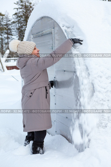Middle age woman trying to clear the snow away from roof of plastic greenhouse at winter season. Стоковое фото, фотограф Кекяляйнен Андрей / Фотобанк Лори