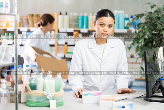 Middle-aged female pharmacist working with computer and other restocking in background. Стоковое фото, фотограф Яков Филимонов / Фотобанк Лори