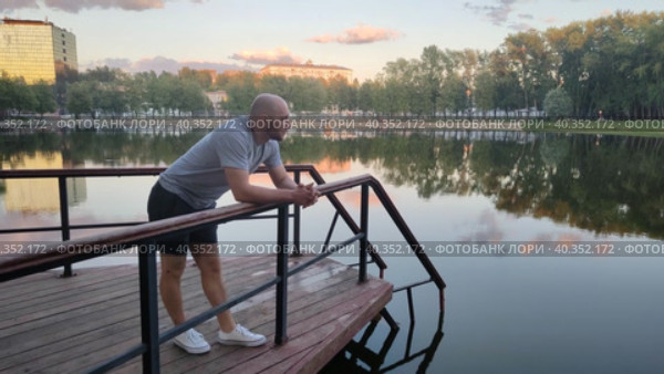 Молодой человек стоит на мостике, смотрит на водную гладь озера в лучах заката. Тихий вечер у пруда в городском парке. Задумчивый парень в спортивной одежде. Активный образ жизни на свежем воздухе. Мужчина на пирсе. A young man is standing on the bridge, looking at the water surface of the lake in the sunset rays. A quiet evening by the pond in the city park. A thoughtful guy in sportswear. An active lifestyle in the fresh air. The man on the pier. Стоковое видео, видеограф Ирина / Фотобанк Лори