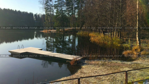 Morning at calm forest lake with frost on earth and wooden pier, cold autumn season. Sunlight from rising sun on the trees. Стоковое видео, видеограф Кекяляйнен Андрей / Фотобанк Лори