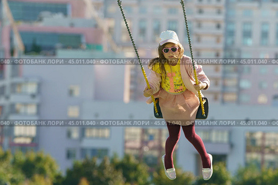 Moscow, Russia - August 20, 2019: Photography of small girl swinging on the sports ground in summer day. She soaring over the city. Concepts of resting, happiness and childhood. Редакционное фото, фотограф YuryKara / Фотобанк Лори