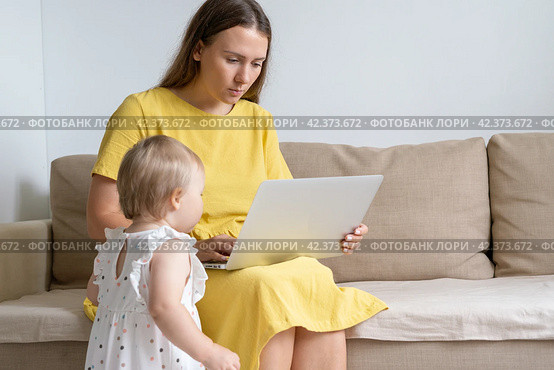 Mother working on laptop during quarantine. Homelife distractions. Curious baby looking at display. Стоковое фото, фотограф Natalia Gdovskaia / Фотобанк Лори