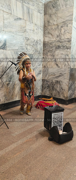 Мужчина индеец в головном уборе роуч с перьями на голове и в кожаной одежде исполняет мелодию на музыкальном инструменте флейте из бамбука в переходе метро арбатская. Редакционное фото, фотограф Светлана Русских / Фотобанк Лори