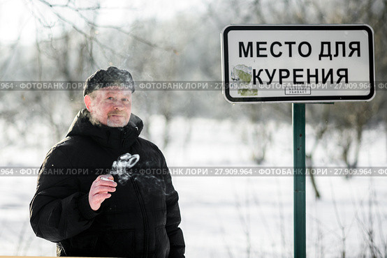 Мужчина курит сигарету возле таблички "Место для курения" зимним днём. Стоковое фото, фотограф Игорь Низов / Фотобанк Лори