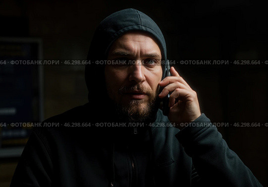 Мужчина в темном капюшоне с сосредоточенным взглядом разговаривает по телефону. Его выражение лица выдает напряженность, что может свидетельствовать о возможном мошенничестве. Стоковое AI-изображение, AI-художник Сергей Никифоров-Гордеев / Фотобанк Лори