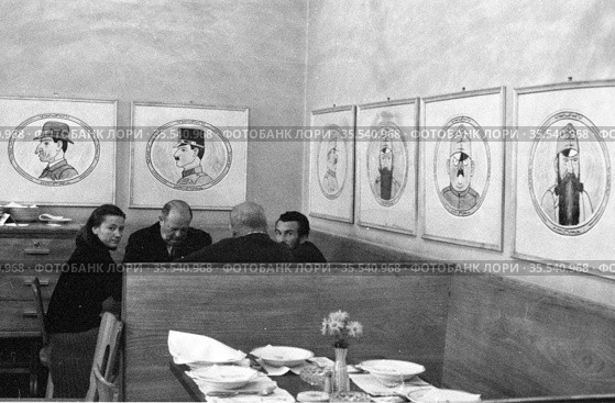 Мужчины и женщина сидят за столом в пивном ресторане "U kalicha", большое количество картин с карикатурными портретами висят на стене, Чехословакия, 1960-е годы. Редакционное фото, фотограф Варфоломей Тетерин / Фотобанк Лори