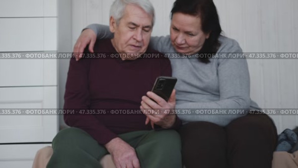 Old couple using a phone together while relaxing in the kitchen at home. Elderly man and woman spending leisure time browsing online with a smartphone. Happy senior married couple typing, learning. Стоковое видео, видеограф Марина Демидюк / Фотобанк Лори