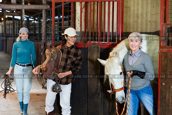 Older woman rancher leading horse out of stall while Asian woman holding saddle. Стоковое фото, фотограф Яков Филимонов / Фотобанк Лори