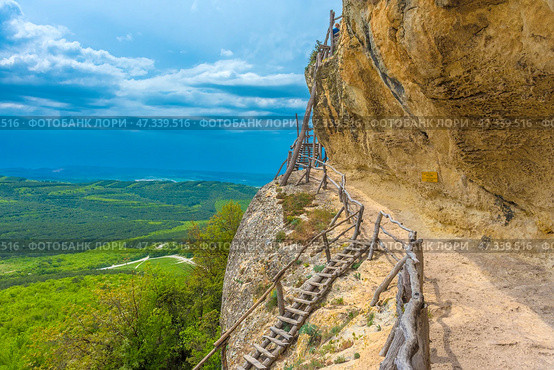One of the picturesque sections of the Chelter-Marmara cave monastery, located in the western part of Crimea, near Bakhchisarai. A narrow path with a wooden railing winds along a rocky cliff leading to a rough staircase made of unprocessed trunks and branches. The cliff seems to hang over the road, and beyond its edge is a bottomless valley and a majestic view of the Crimean foothills.This place is filled with the spirit of hermitage, peace and the grandeur of nature. A sign on the wall of the cliff warns of the height and calls for attention, reminding of the harsh but beautiful conditions in which the monks lived. Everything here - both the architecture and the rock landscape - is like a living testimony to time and the power of faith. Стоковое фото, фотограф Анна Кривицкая / Фотобанк Лори