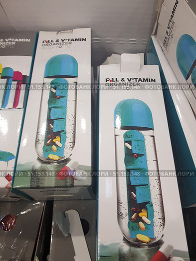 Packaging of a multifunctional water bottle with a built-in organizer for pills and vitamins. The box shows a transparent bottle with a blue lid and a built-in container with compartments for capsules and pills for each day of the week. The packaging demonstrates a practical solution for those who take vitamins or medications daily. Such an accessory combines the convenience of storing medications and the ability to always have water on hand to take them. In the foreground are capsules and tablets, emphasizing the purpose of the product. (2019 год). Редакционное фото, фотограф Анна Кривицкая / Фотобанк Лори