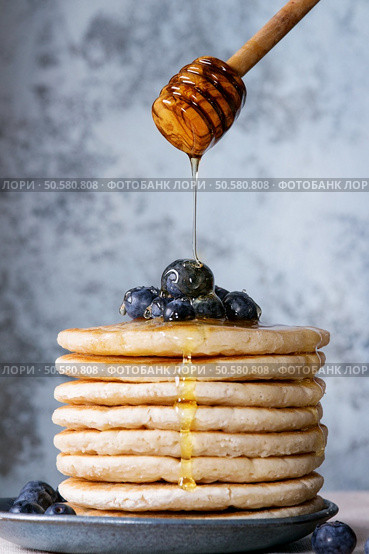 Pancakes with fresh blueberries in blue ceramic plate, and flowing honey from wooden honey dipper, served on white tablecloth with blue textured background.. Pancakes with fresh berries. Стоковое фото, агентство Ingram Publishing / Фотобанк Лори