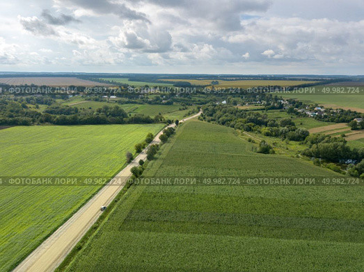 Panoramic view from drone to the countryside with a country buildings, dirt road and agricultural fields against cloudy sky in the summer at sunset.. Bird’s eye view from drone to a rural landscape with a village, dirt road and agricultural fields of planted crops at cloude background. Стоковое фото, агентство Ingram Publishing / Фотобанк Лори