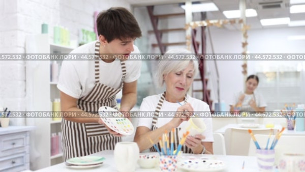 Passionate elderly female teacher offering guidance to interested young guy painting ceramic mug in pottery class. Creative hobby concept. Стоковое видео, видеограф Яков Филимонов / Фотобанк Лори