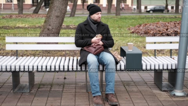 Pensive hipster drinking coffee sitting on a bench in the urban park. Handsome man wearing black coat eating street food, hot dog with paper cup in hand in the city. Stylish bearded middle-aged male. Стоковое видео, видеограф Марина Демидюк / Фотобанк Лори