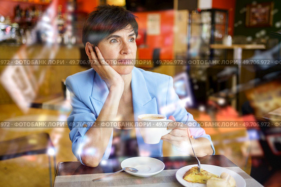 Pondering woman in stylish blue jacket sitting at table with cup of hot drink and looking away during breakfast in cafe. Wistful lady drinking coffee in cafeteria. Стоковое фото, агентство Ingram Publishing / Фотобанк Лори