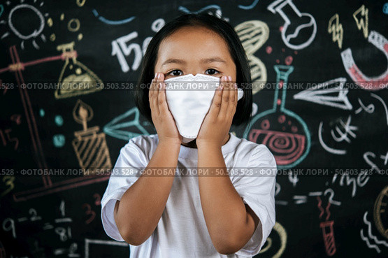 Portrait of a little girl wearing eyeglasses and facemask at school to avoid the spread of coronavirus, new normal with schoolchild,  blure blackboard in background. Стоковое фото, агентство Ingram Publishing / Фотобанк Лори