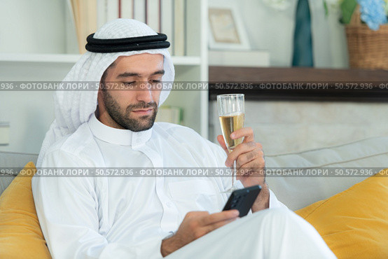 Portrait of a smiling Arabian man holding a glass of champagne and using mobile phone at home. Стоковое фото, агентство Ingram Publishing / Фотобанк Лори