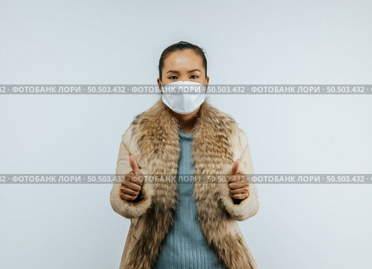 Portrait of Asian woman with two thumbs up sign gesture, putting on protective mask on face for protection against coronavirus, wearing a fur coat standing over background in studio with copy space. Стоковое фото, агентство Ingram Publishing / Фотобанк Лори