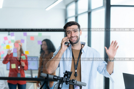Portrait of smiling businessman talking on mobile phone while colleague using scooter in office. Стоковое фото, агентство Ingram Publishing / Фотобанк Лори