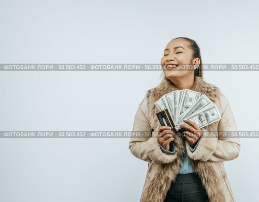 Portrait of successful Asian woman with a fur coat holding money and card, smiling and closing eyes, standing over background in studio with copy space. Стоковое фото, агентство Ingram Publishing / Фотобанк Лори