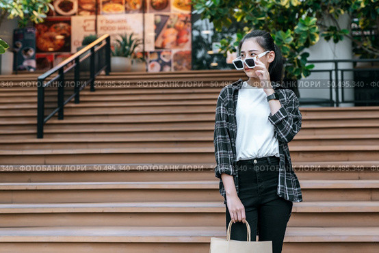 Portrait of young Asian woman wearing eyeglasses going down stairs with shopping paper bag. she smile and looking at camera,  Lifestyle and shopping concept, Shopping selection. Стоковое фото, агентство Ingram Publishing / Фотобанк Лори