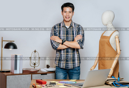 Portrait young Asian fashion designer or tailor working with laptop computer, standing with poud, full of tailoring tools with mannequin and equipment on desk in studio. Стоковое фото, агентство Ingram Publishing / Фотобанк Лори