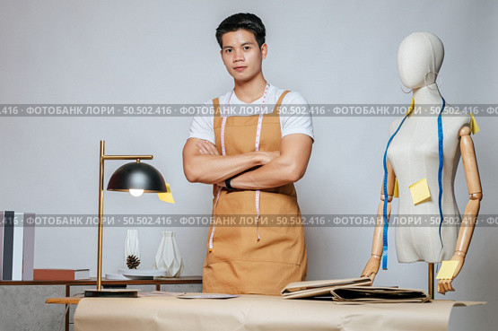 Portrait Young male fashion designer working with fabric and clothing sketches at the studio at home, full of tailoring tools, model and equipment on table. Стоковое фото, агентство Ingram Publishing / Фотобанк Лори