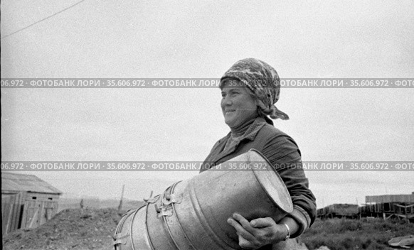 Портрет зрелой женщины с платком на голове держащая в руках несколько алюминиевых вёдер, деревянные постройки на заднем плане, северное птицеводство, Лорино, Чукотка, 1970. Редакционное фото, фотограф Варфоломей Тетерин / Фотобанк Лори