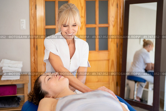 Positive female osteopath in uniform manipulating head of patient lying on couch during psychotherapy session in physical therapy clinic. Smiling physiotherapist treating patient in hospital room. Стоковое фото, агентство Ingram Publishing / Фотобанк Лори