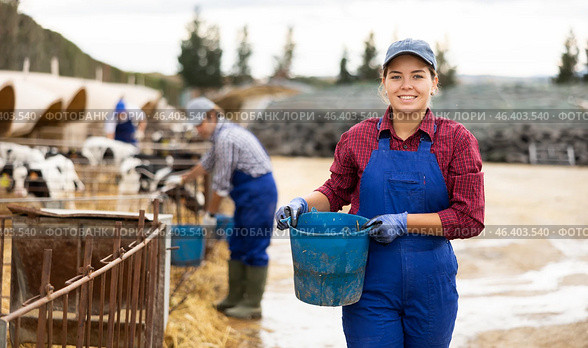 Positive woman farmer worker in uniform preparing water in buckets for taking care of calves at livestock farm. Стоковое фото, фотограф Яков Филимонов / Фотобанк Лори