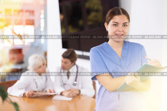 positive young medical female colleague standing in meeting room and employees are talking behind her at table. Стоковое фото, фотограф Яков Филимонов / Фотобанк Лори