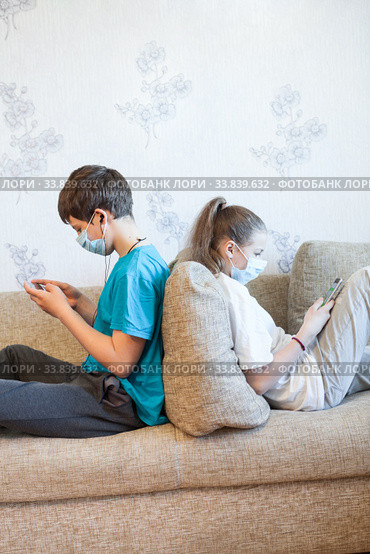 Pre-teen children gaming online using cellphones while sitting at home, kids wearing facial masks, sitting back to back on couch. Стоковое фото, фотограф Кекяляйнен Андрей / Фотобанк Лори