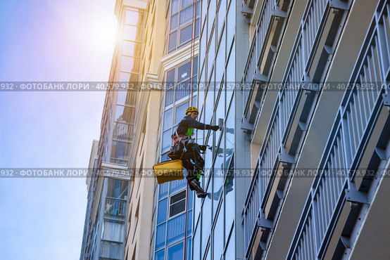 Professional industrial mountaineer worker washing exterior facade glazing, hangs over building, view from below. Rope access laborer climbing on wall house. Urban works concept. Copy ad text space. Стоковое фото, фотограф Aleksandr Golubev / Фотобанк Лори