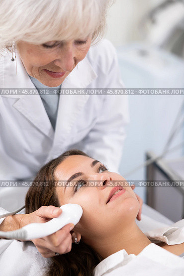 Professional mature female cosmetologist doing facial skin ultrasound cavitation for flawless woman face laying in beauty salon. Стоковое фото, фотограф Яков Филимонов / Фотобанк Лори