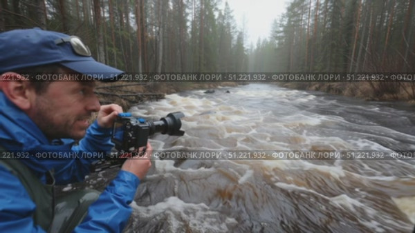Professional photographer documenting wild river dynamics in boreal forest, wearing waterproof gear near rushing stream during overcast weather conditions. Стоковое AI-видео, аниматор Юрий Поздников / Фотобанк Лори