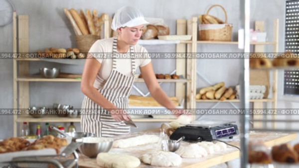 Professional young female baker in white uniform carefully slicing and weighting dough on flour-covered worktable in cozy bakery filled with freshly baked bread and baking tools. Стоковое видео, видеограф Яков Филимонов / Фотобанк Лори