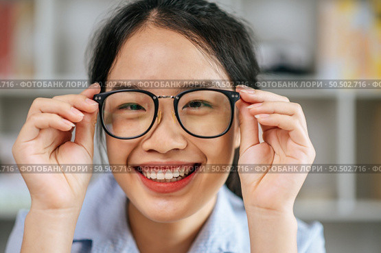 Protrait young asian woman happy positive smile and use hand touch eyeglasses and looking at camera at living room. Стоковое фото, агентство Ingram Publishing / Фотобанк Лори