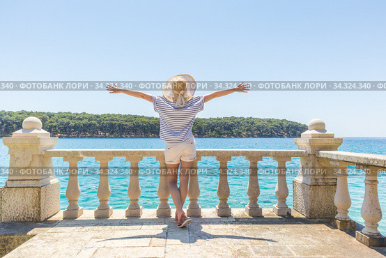 Rear view of happy woman on vacation, wearing straw summer hat ,standing on luxury elegant old stone balcony of coastal villa, relaxing, arms rised to the sun, looking at blue Adriatic sea. Стоковое фото, фотограф Matej Kastelic / Фотобанк Лори