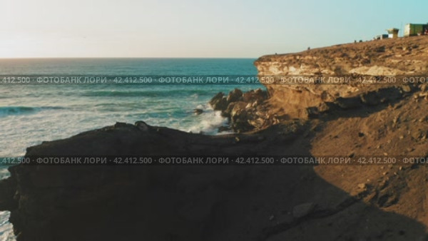 Rocky coastline alongside cliff, blue Atlantic Ocean water reflect at sunset. Nature landscape. Golden sandstone bluff beach. Tourists meet dusk. Travel destination. Fuerteventura Island, Canaries. (2022 год). Стоковое видео, видеограф Igor Tichonow / Фотобанк Лори