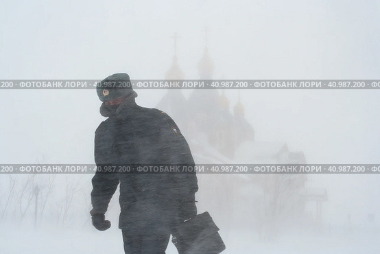 Российский полицейский идет по городу во время сильной метели. Ветреная и снежная погода. Снежная буря и пурга в городе. Вдали собор. Анадырь, Чукотка, Россия. Редакционное фото, фотограф Андрей Степанов / Фотобанк Лори