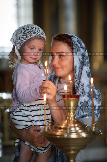 Russian beautiful woman in a scarf and with red hair holding a little girl and lights a candle in front of an icon in the Russian Orthodox Church. Стоковое фото, фотограф Арзюков Вадим Николаевич / Фотобанк Лори